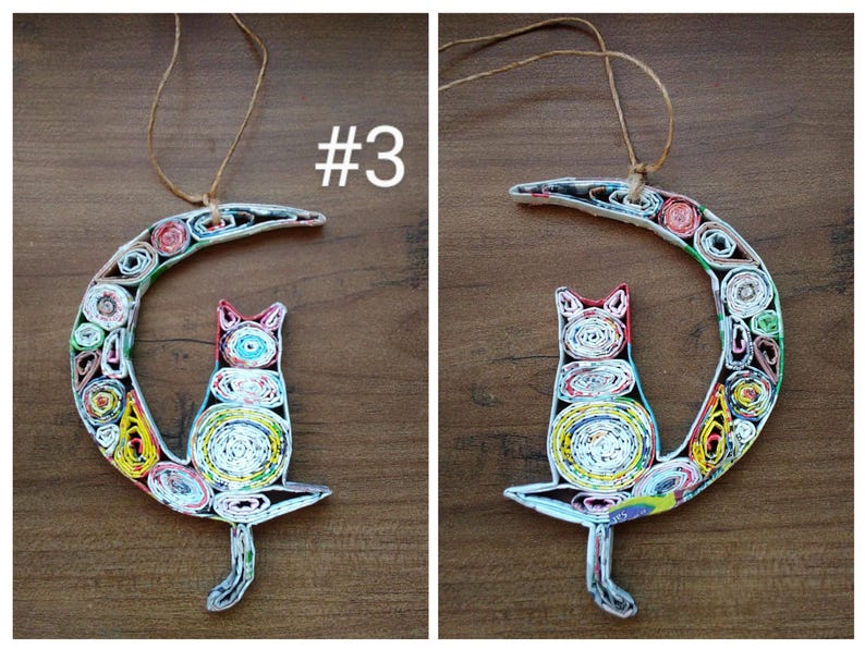 May include: A colorful paper mache cat sitting on a crescent moon. The cat and moon are made of rolled up paper and have a multi-colored pattern. The cat is white with red, blue, and yellow accents. The moon is white with green, yellow, red, and blue accents. #3