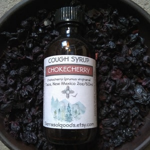 May include: A dark brown glass bottle with a black cap, filled with a dark liquid. The label reads "Cough Syrup Chokecherry Chokecherry (prunus virginiana) Taos, New Mexico 2oz/60ml" with a stylized image of a mountain range and a bird. The bottle is sitting on a bed of dried chokecherries.