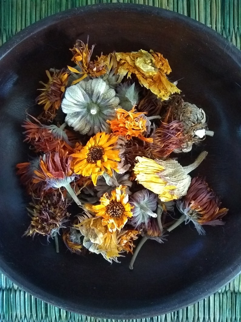 May include: A collection of dried calendula flowers in shades of orange, yellow, and brown, clustered together inside a dark brown, circular wooden bowl. The dried petals and flower heads show varied textures and natural, muted earth tones.