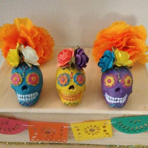 May include: Three colorful sugar skull decorations with flower accents. The skulls are painted in blue, yellow, and purple with white accents. The flowers are orange, yellow, white, and blue.