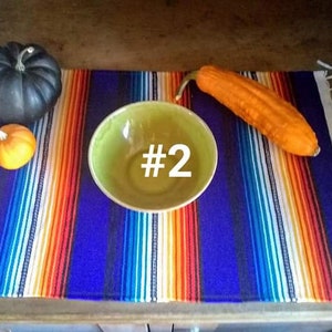 May include: A colorful striped textile with a blue background, orange, white, and yellow stripes. A green bowl with the number 2 in white, a dark gray pumpkin, a small orange pumpkin, and a long orange gourd are on the textile.