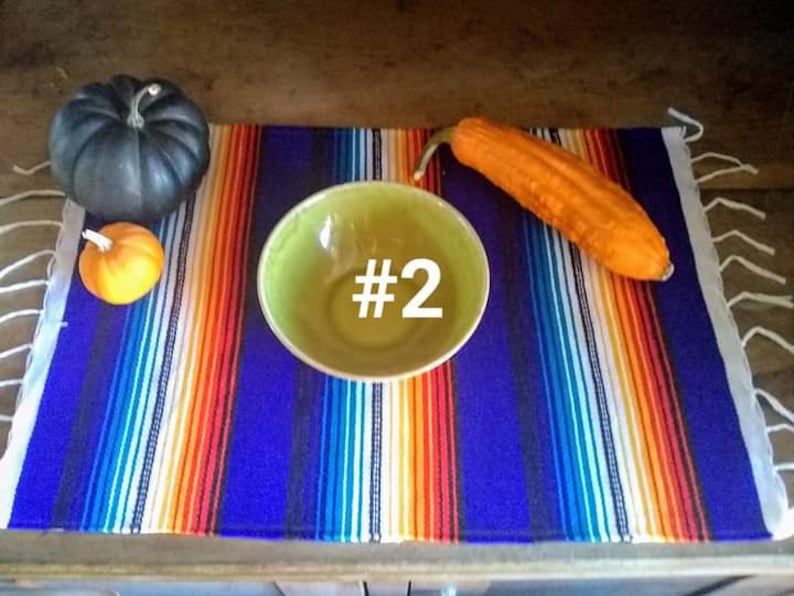 May include: A colorful striped textile with a blue background, orange, white, and yellow stripes. A green bowl with the number 2 in white, a dark gray pumpkin, a small orange pumpkin, and a long orange gourd are on the textile.