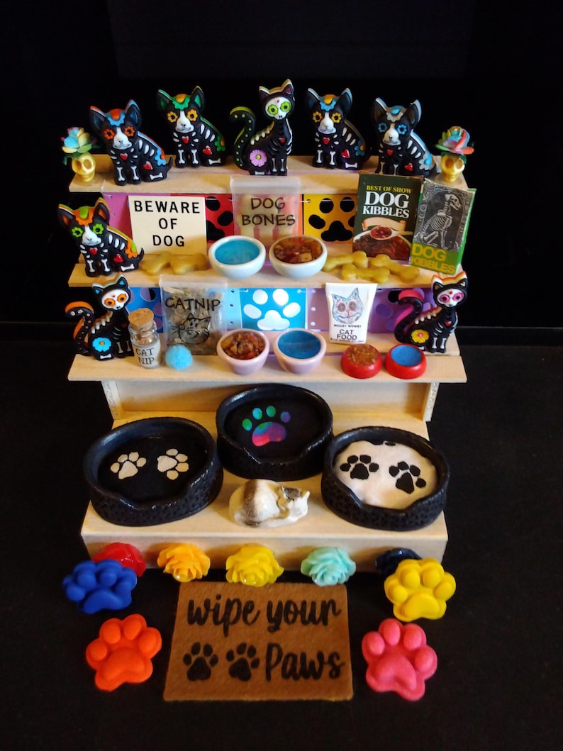 May include: A multi-tiered display of pet-themed items. The top shelf features Day of the Dead cat figurines, bowls, and signs that read "Beware of Dog" and "Dog Bones." The middle shelf has cat food and catnip. The bottom shelf has paw-shaped items.