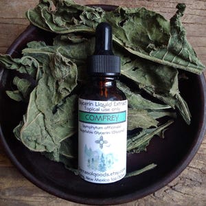 May include: A small brown glass bottle with a dropper top, labeled "Glycerin Liquid Extract Topical use only COMFREY (symphytum officinale) Vegetable Glycerin: (Glycerol + terrasolgoods.etsy.com New Mexico 1oz/30ml". The bottle is sitting on a brown wooden surface with dried green leaves.