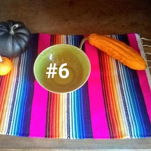 May include: A vibrant, striped textile with a bright pink base, adorned with a green bowl with the number 6, a black pumpkin, a small orange pumpkin, and an orange gourd. The textile has white fringe.