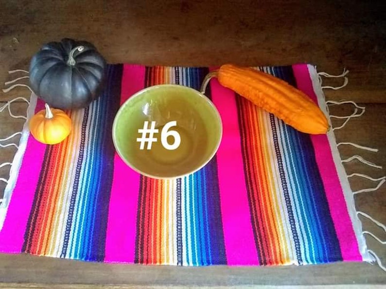 May include: A vibrant, striped textile with a bright pink base, adorned with a green bowl with the number 6, a black pumpkin, a small orange pumpkin, and an orange gourd. The textile has white fringe.