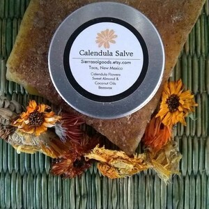 May include: A round metal tin of Calendula Salve with a white label featuring the text Calendula Salve, Sierrasolgoods.etsy.com, Taos, New Mexico, Calendula Flowers, Sweet Almond & Coconut Oils, Beeswax. The tin rests on a textured brown block.
