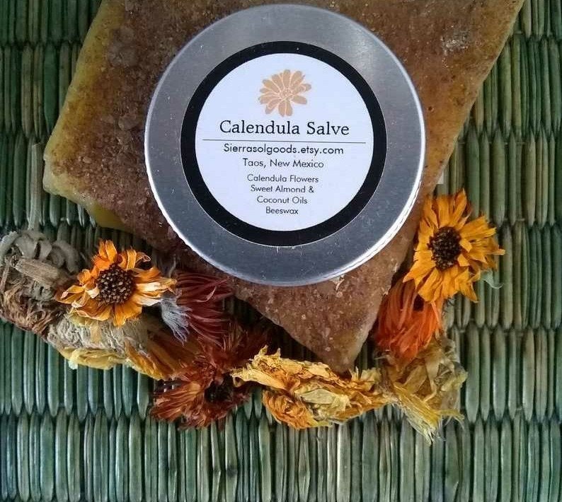 May include: A round metal tin of Calendula Salve with a white label featuring the text Calendula Salve, Sierrasolgoods.etsy.com, Taos, New Mexico, Calendula Flowers, Sweet Almond & Coconut Oils, Beeswax. The tin rests on a textured brown block.