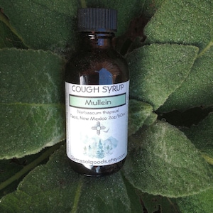 K&ouml;nnte beinhalten: Eine braune Glasflasche mit schwarzem Verschluss und einem wei&szlig;en Etikett mit der Aufschrift "Cough Syrup Mullein (Verbascum thapsus) Taos, New Mexico 2oz/60ml" mit einem stilisierten Berg- und Kreuzdesign. Die Flasche steht auf einer gr&uuml;nen, bebl&auml;tterten Oberfl&auml;che.