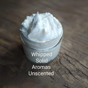 May include: A white whipped solid body butter in a glass jar on a wooden surface. The jar has a label that reads "Whipped Solid Aromas Unscented".