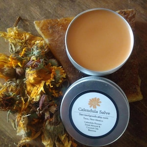May include: A round silver metal tin containing orange calendula salve, with a matching lid featuring a white label that reads Calendula Salve, Sierrasolgoods.etsy.com, Taos, New Mexico, Calendula Flowers, Sweet Almond & Coconut Oils, Beeswax.