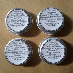 Puede incluir: Cuatro latas plateadas con etiquetas blancas que contienen diferentes b&aacute;lsamos herbales. Cada etiqueta enumera los ingredientes, incluyendo aceite de almendras dulces, aceite de coco, cera de abejas y el nombre latino de la hierba. Las etiquetas tambi&eacute;n indican "Reci&eacute;n hecho".
