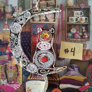May include: A paper mache cat ornament in the shape of a crescent moon. The cat is made of rolled up paper and has a red, yellow, and white pattern. The ornament is hanging from a string and has a tag that says "#4".