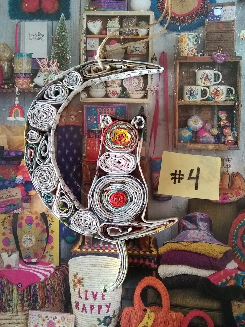 May include: A paper mache cat ornament in the shape of a crescent moon. The cat is made of rolled up paper and has a red, yellow, and white pattern. The ornament is hanging from a string and has a tag that says "#4".