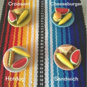 May include: Four miniature plates with different food items on each. The plates are on a colorful striped blanket. The plates are labeled "Croissant", "Cheeseburger", "Hotdog", and "Sandwich".