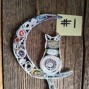 May include: A handmade paper cat ornament, curled up on a crescent moon, made from rolled up colorful magazine pages. The ornament is hanging from a string and has a yellow sticky note with the number "#1" written on it.
