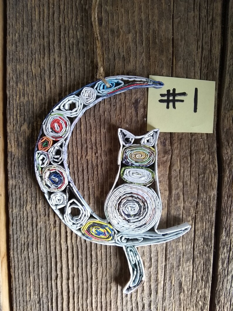 May include: A handmade paper cat ornament, curled up on a crescent moon, made from rolled up colorful magazine pages. The ornament is hanging from a string and has a yellow sticky note with the number "#1" written on it.