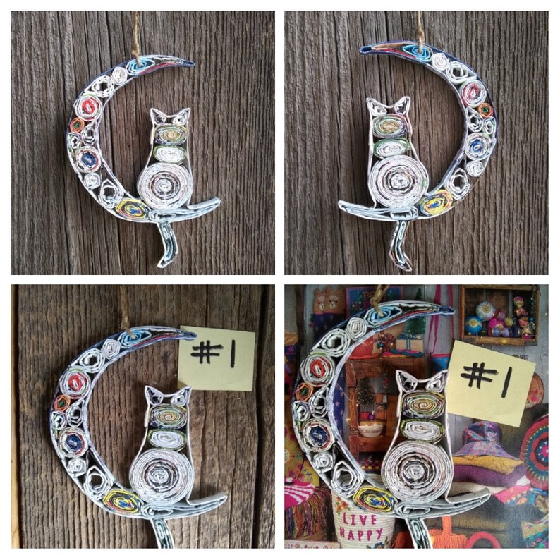 May include: A colorful paper mache cat sitting on a crescent moon. The cat and moon are made of rolled up paper and have a multi-colored pattern. The background is a wooden surface.