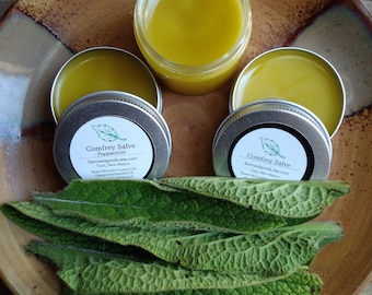 May include: Two small round tins of Comfrey Salve, one with peppermint and one without, on a brown plate with green leaves. The tins have white labels with black text that reads "Comfrey Salve" and "Sierragoldgoods.etsy.com".