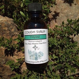May include: A dark glass bottle of cough syrup with a black cap, labeled "COUGH SYRUP THYME." The label includes the text "(thymus vulgaris)" and "Taos, New Mexico 2oz/60ml." The bottle is surrounded by green foliage and rests on a rock.
