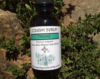 Könnte beinhalten: Eine dunkle Glasflasche Hustensaft mit schwarzem Verschluss, beschriftet mit "COUGH SYRUP THYME". Das Etikett enthält den Text "(thymus vulgaris)" und "Taos, New Mexico 60ml". Die Flasche ist von grünem Laub umgeben und liegt auf einem Felsen.