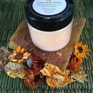 May include: A round, clear plastic jar with a black lid containing Calendula Salve. The label lists Calendula Flowers, Sweet Almond & Coconut Oils, and Beeswax. The jar rests on a rectangular block of amber-colored wax surrounded by dried orange flowers.