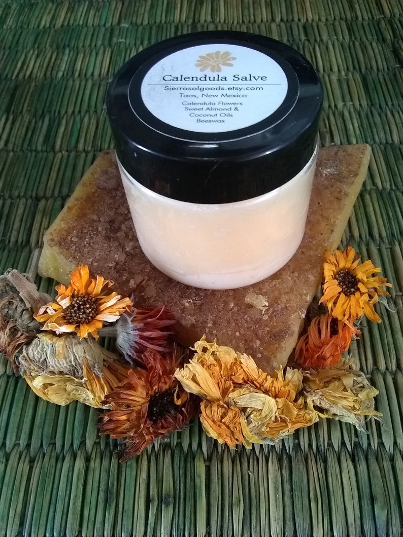 May include: A round, clear plastic jar with a black lid containing Calendula Salve. The label lists Calendula Flowers, Sweet Almond & Coconut Oils, and Beeswax. The jar rests on a rectangular block of amber-colored wax surrounded by dried orange flowers.