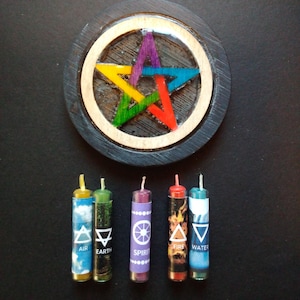 May include: A wooden pentacle with a colorful star design and five small pillar candles. Each candle has a different color and symbol, including Air, Earth, Spirit, Fire, and Water. The candles are cylindrical and have a white wick.