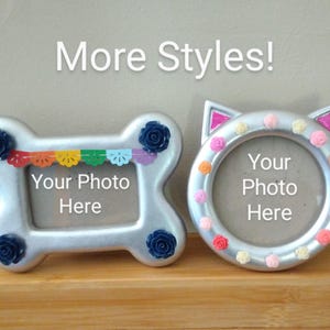 May include: Two silver picture frames with the text 'More Styles!' above them. The left frame is bone-shaped with dark blue roses and a colorful banner. The right frame is cat-shaped with small pink, orange, and white roses. Both have 'Your Photo Here' text.