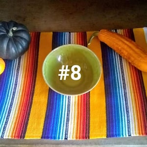 May include: A still life arrangement featuring a green bowl with the number 8, a colorful striped textile, and gourds. The gourds include a dark blue pumpkin and a small orange gourd. An orange squash is also present.