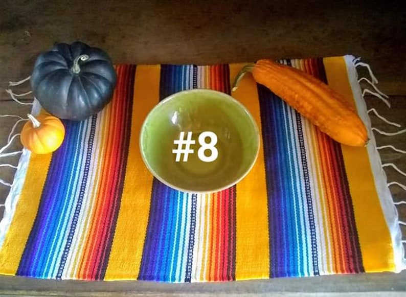 May include: A still life arrangement featuring a green bowl with the number 8, a colorful striped textile, and gourds. The gourds include a dark blue pumpkin and a small orange gourd. An orange squash is also present.