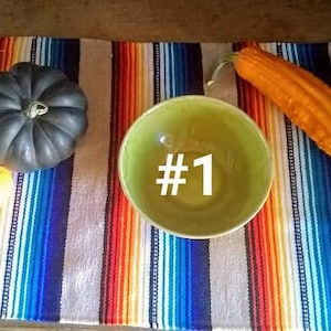 May include: A still life arrangement featuring a green bowl with the number 1, a small orange pumpkin, a dark gray pumpkin, and a long orange gourd. The items are arranged on a colorful striped textile.