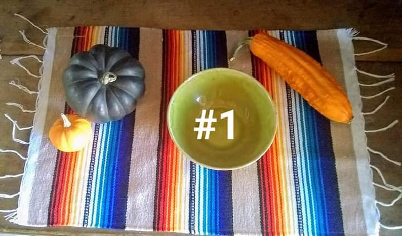 May include: A still life arrangement featuring a green bowl with the number 1, a small orange pumpkin, a dark gray pumpkin, and a long orange gourd. The items are arranged on a colorful striped textile.