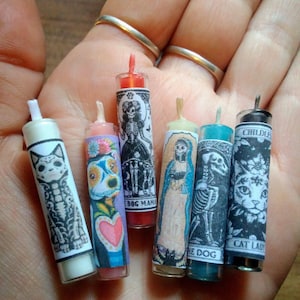 May include: Six miniature cylindrical candles featuring various graphic labels. The labels include illustrations of a cat skeleton, a colorful dog, a skeleton figure labeled BOG MAMA, a robed figure, a dog skeleton labeled THE DOG, and a cat labeled CAT LADY.