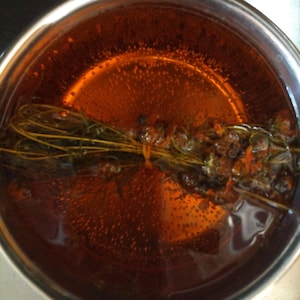 May include: A stainless steel pot filled with a clear amber liquid. Submerged in the liquid is a bundle of green herbs tied with a thin orange string. Small bubbles are visible throughout the liquid, concentrated around the herb bundle.