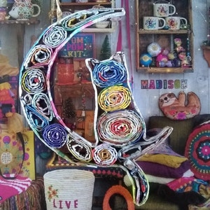 May include: A colorful cat and crescent moon sculpture made from rolled up paper. The sculpture is decorated with a variety of colors and patterns. The cat is sitting on the crescent moon and has a playful expression.