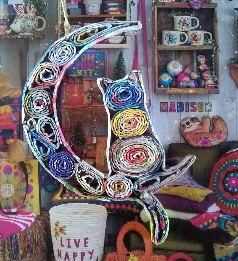 May include: A colorful cat and crescent moon sculpture made from rolled up paper. The sculpture is decorated with a variety of colors and patterns. The cat is sitting on the crescent moon and has a playful expression.