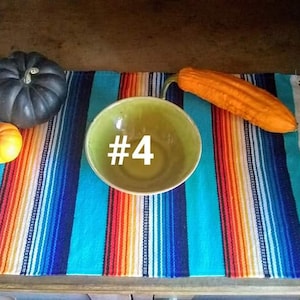 May include: A still life arrangement featuring a green bowl with the number 4, a dark gray pumpkin, a small orange pumpkin, and an orange gourd. The items are placed on a colorful striped textile with fringe.