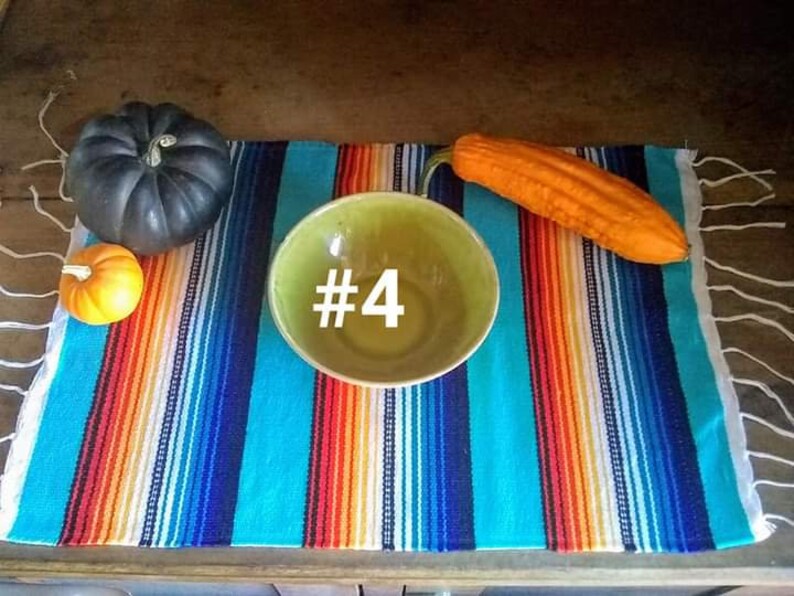 May include: A still life arrangement featuring a green bowl with the number 4, a dark gray pumpkin, a small orange pumpkin, and an orange gourd. The items are placed on a colorful striped textile with fringe.
