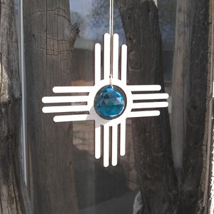May include: White wooden cutout of the Zia symbol, a traditional symbol of the Pueblo people, with a blue crystal in the center. The symbol is hanging from a string.