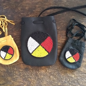 May include: Three leather pouches with drawstrings, each featuring a beaded Medicine Wheel design in black, white, yellow, and red. The pouches are different sizes, with the largest being black, the medium being tan, and the smallest being black.