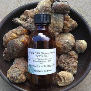 May include: A 2 ounce amber glass bottle of Piñon Sap body oil with a white label. The label reads "Piñon Sap (Trementina) Body Oil Sweet Almond-Coconut Olive Oils-Piñon Sap 2oz Sierrasolgoods.etsy.com Taos New Mexico". The bottle is surrounded by pieces of brown, yellow, and white resin.