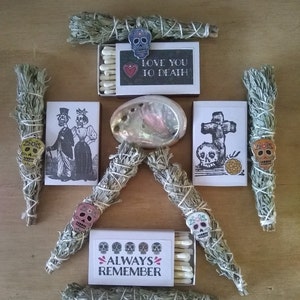 May include: An assortment of Day of the Dead themed items, including bundles of sage, matchboxes with skull and skeleton illustrations, and an abalone shell. The matchboxes have text that reads "LOVE YOU TO DEATH" and "ALWAYS REMEMBER".