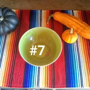 May include: A still life arrangement featuring a dark green gourd, a long orange squash, and a small orange pumpkin. A green bowl with the number 7 in white is centered on a colorful striped textile.