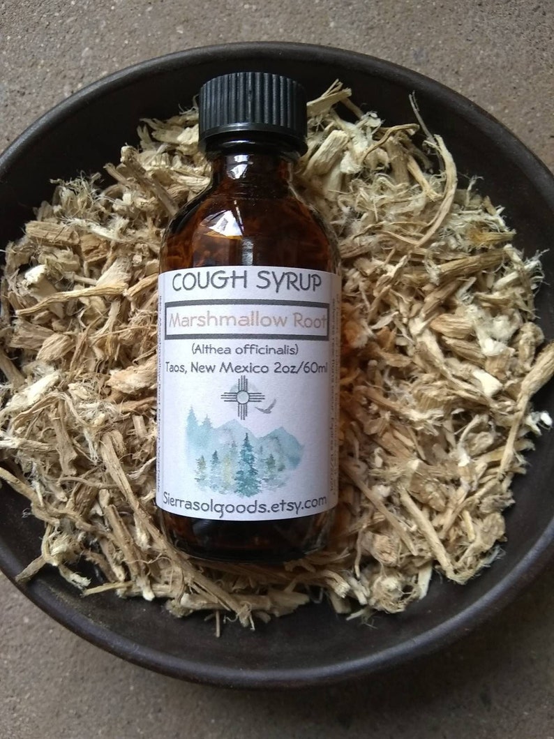 May include: A glass bottle of cough syrup with a white label that reads "COUGH SYRUP Marshmallow Root (Althea officinalis) Taos, New Mexico 2oz/60ml" with a stylized image of a mountain range and a sun symbol. The bottle is sitting on a bed of dried, light brown, shredded plant material. The text "Sierrasolgoods.etsy.com" is printed on the label.