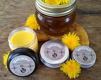 May include: An assortment of dandelion salve products, including a jar filled with yellow dandelion flowers, and several small tins and jars of salve. The products are arranged on a wooden surface, with additional yellow dandelion flowers scattered around.