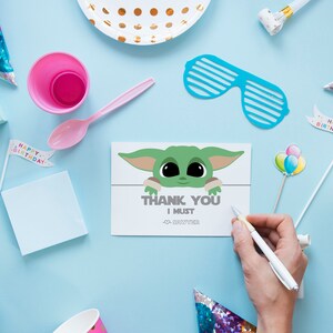 May include: A thank you card with a cartoon image of a green alien character with big eyes. The card says "Thank You I Must" and has a small alien character in the corner. The card is surrounded by party decorations, including a pink plate, a pink cup, a blue party hat, and a white cup.