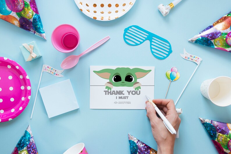 May include: A thank you card with a cartoon image of a green alien character with big eyes. The card says "Thank You I Must" and has a small alien character in the corner. The card is surrounded by party decorations, including a pink plate, a pink cup, a blue party hat, and a white cup.