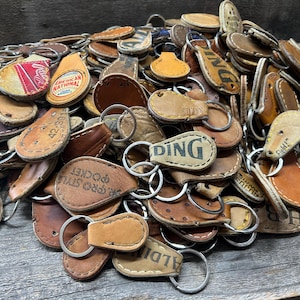 May include: A large pile of leather keychains with various designs and text, including "DING", "PRO STYLE POCKET", and "AMERICAN NATIONAL".