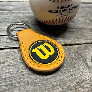 May include: A tan leather keychain with a yellow and black embroidered "W" logo. The keychain is attached to a silver key ring.